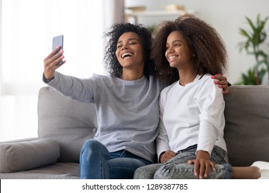 Happy Millennial Black Mom Make Selfie On Smartphone With Teenage Daughter, Smiling Mother And Teen Child Shot Self Portrait Picture On Cell Phone At Home. Concept Of Parent And Children Relationships