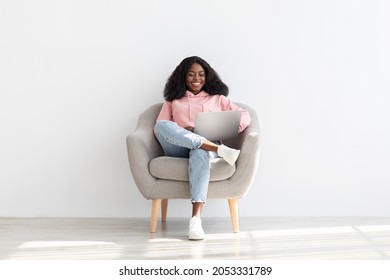 Happy Millennial Black Woman In Casual Outfit Independent Contractor Sitting In Stylish Arm Chair By White Empty Wall, Using Modern Laptop, Working From Home, Copy Space, Full Size Shot