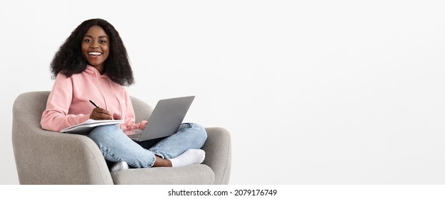 Happy Millennial Black Woman In Casual Outfit Independent Contractor Sitting In Stylish Arm Chair By White Empty Wall, Using Modern Laptop, Taking Notes, Working From Home, Copy Space, Panorama