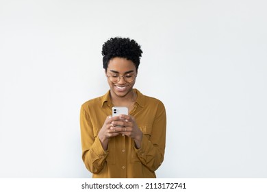 Happy Millennial Black Woman In Casual Glasses Browsing Internet On Mobile Phone, Reading, Typing Text Message, Chatting Online, Smiling, Laughing, Using Banking App On Smartphone. Isolated Portrait