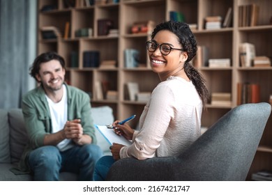 Happy millennial black woman psychologist in glasses look at camera, consulting smiling european man in office clinic interior. Therapy, mental problems, psychologist support and professional help - Shutterstock ID 2167421747