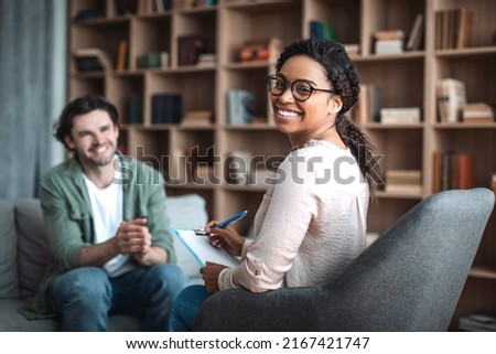 Happy millennial black woman psychologist in glasses look at camera, consulting smiling european man in office clinic interior. Therapy, mental problems, psychologist support and professional help