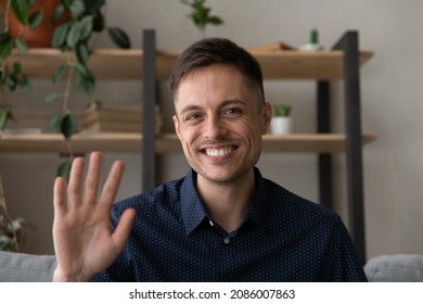 Happy millennial business man in casual making video call from home couch, looking at camera, smiling, waving hand hello, attending online chat, virtual conference. Video call head shot portrait