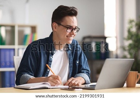 Happy millennial businessman taking notes while working with laptop in office, handsome young male entrepreneur looking at computer screen and smiling, writing down information from internet