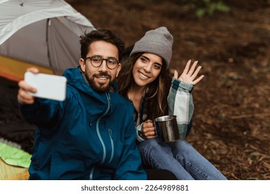 Happy millennial caucasian couple in jackets resting in cold forest, enjoy adventure, sit in tent, taking selfie on phone, waving hand and drink coffee outdoor. Travel blog, vacation and camping - Powered by Shutterstock