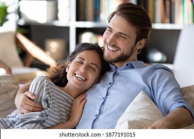 Happy Millennial Caucasian Couple Relax On Couch At Home Hugging Dreaming Visualizing Together. Smiling Overjoyed Young Man And Woman Rest Embrace On Sofa Enjoy Romantic Family Time On Weekend.