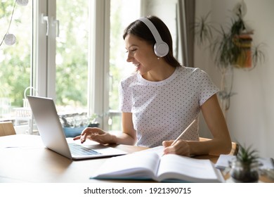 Happy millennial Caucasian female student in headphones look at laptop screen study distant from home. Smiling young woman in earphones make notes watch webinar on computer, take web course. - Shutterstock ID 1921390715