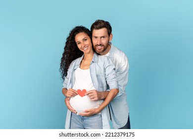 Happy Millennial Caucasian Man With Beard Hugging Pregnant Arabic Woman, Holding Heart On Belly, Isolated On Blue Background, Studio. Couple Enjoy Romance, Relationship And Love Expecting Baby, Ad