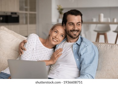 Happy Millennial Caucasian Man And Woman Spouses Use Computer Look In Distance Dreaming Imagining. Smiling Young Couple Relax On Sofa At Home Visualize Happy Future Together. Vision Concept.