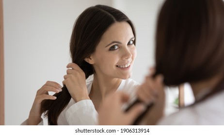 Happy Millennial Caucasian Woman Look In Mirror Satisfied With Healthy Thick Hair After Salon Treatment Or Restoration Mask. Smiling Young Female Do Hairstyle Show Beauty Cosmetic Product Results.