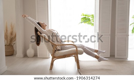 Happy millennial Caucasian woman sit in cozy chair in design home stretch relax breathe fresh air. Smiling young female renter or tenant rest in armchair relieve negative emotions enjoying weekend.