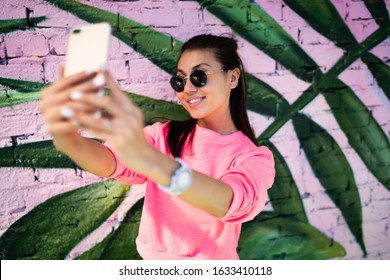 Happy millennial content maker shooting influence video vlog near photo zone enjoying networking lifestyle, cheerful Asian hipster girl in sunglasses smiling at smartphone camera for selfie image