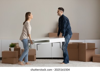 Happy millennial couple carrying coffee table together to unfurnished living room furnish new house with modern comfort furniture. Bank loan, moving, interior design items shop advertisement concept