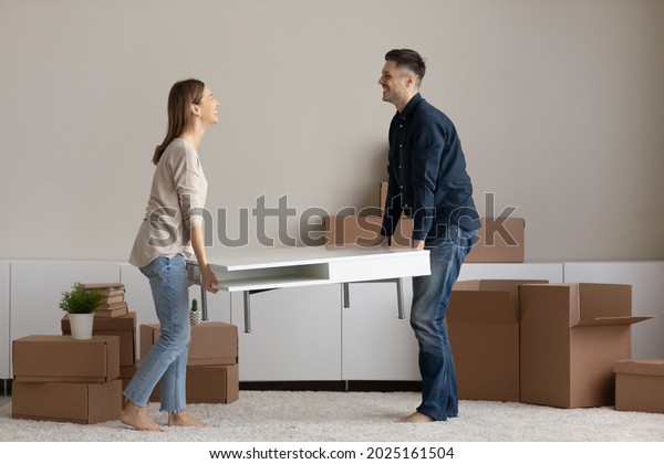 Happy millennial couple carrying coffee table\
together to unfurnished living room furnish new house with modern\
comfort furniture. Bank loan, moving, interior design items shop\
advertisement concept