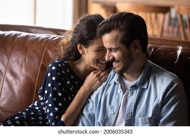 Happy millennial couple enjoying being together in new house, apartment, excited about good news, talking, hugging, laughing, smiling while sitting on luxury leather couch. Love, relationship concept