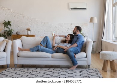 Happy millennial couple of homeowners enjoying cool conditioned air, resting on couch together, using remote control for AC, cooling domestic equipment start. Home appliance concept - Shutterstock ID 2184585771