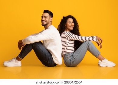 Happy Millennial Couple. Portrait Of Cheerful Young Arab Man And Woman Sitting Back To Back And Smiling At Camera, Joyful Middle Eastern Spouses Posing Over Yellow Background In Studio, Free Space