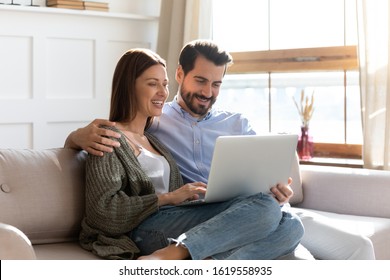 Happy Millennial Couple Sit Relax On Couch In Living Room Watching Video On Laptop Together, Smiling Young Husband And Wife Rest On Sofa At Home Browsing Internet Using Modern Computer Device