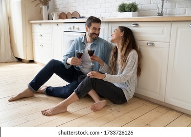 Happy millennial couple sit relax on wooden floor in kitchen drinking red wine talking chatting, smiling young husband and wife rest at home enjoy romantic date on family weekend together - Powered by Shutterstock