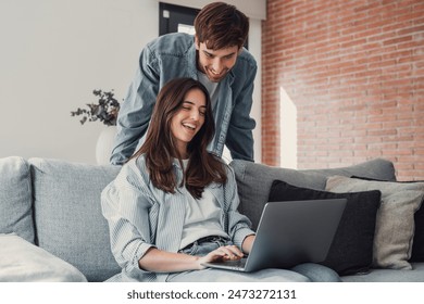 Happy millennial couple sit relax on couch in living room, smiling young husband standing from back and wife rest on sofa at home browsing Internet using modern computer device - Powered by Shutterstock