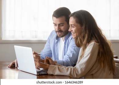 Happy millennial couple sit at table at home browsing web on laptop shopping online together, smiling young husband and wife work on computer at desk pay household bills or taxes in internet banking