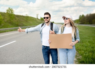 Happy Millennial Couple Standing Along Highway With Empty Sign, Stopping Car, Having Road Trip, Mockup For Design. Wanderlust, Autostop Journey, Hitchhiking Together, Summer Freedom Concept