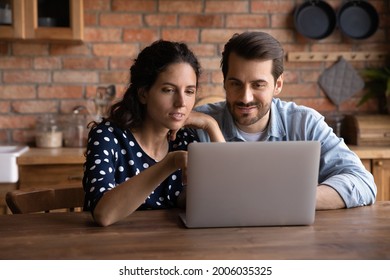 Happy Millennial Couple Using Laptop In Kitchen, Watching Show Together, Making Video Call, Paying Bills. Young Husband And Wife Using Online App On Computer At Home, Looking At Screen, Smiling