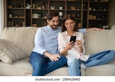 Happy millennial couple using online shopping app, internet service, making video phone call, sharing cellphone, watching media content together, talking, resting on sofa with digital device at home