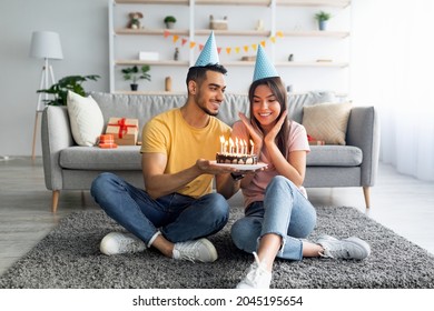 Happy millennial couple wearing party hats, celebrating birthday with festive cake at home, full length. Cheerful Arab guy greeting excited girlfriend with b-day, enjoying holiday in living room