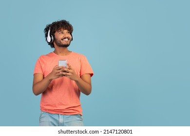 Happy millennial eastern guy in pink t-shirt using brand new wireless stereo headset and smartphone, listening to music over blue studio background, looking at copy space and smiling, panorama - Powered by Shutterstock