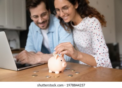 Happy Millennial Family Couple Putting Coins In Piggybank, Planning Vacation Or Investments Together, Saving Money For Life Insurance, Managing Future Expenditures Together Using Computer Apps.