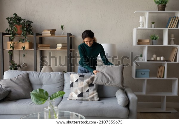Happy millennial generation positive beautiful\
young housewife cleaning apartment, arranging stuff in cozy modern\
living room, put things on right places, feeling satisfied with\
comfort tidy house.