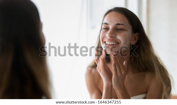 Happy\
millennial girl look in mirror after shower in home bath do facial\
scrub mask massage skin, smiling young woman perform daily face\
morning routine or beauty procedures in\
bathroom