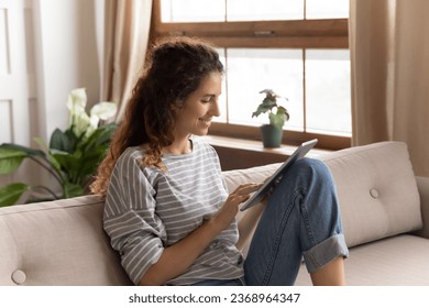 Happy millennial girl sit on sofa in living room browsing wireless Internet on modern tablet gadget, smiling young woman relax on couch at home reading book or surfing web on Pad, technology concept - Powered by Shutterstock