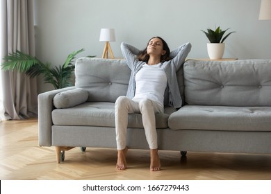 Happy millennial girl sit relax on comfortable couch in living room hands over head, peaceful young woman rest on cozy sofa at home take nap dream with closed breathe fresh air, stress free concept