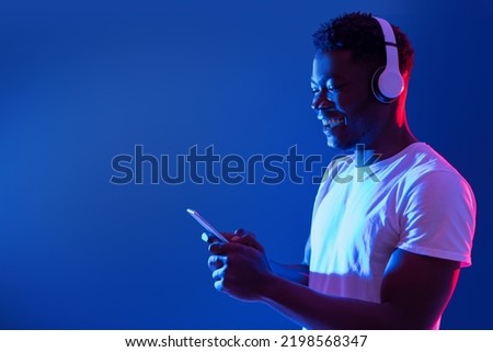 Happy millennial guy in wireless headphones using smartphone in blue neon light, copy space. Handsome young African American man checking new mobile music app online