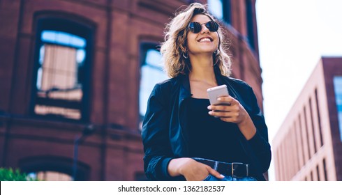 Happy millennial hipster girl in sunglasses laughing on urban setting during sunny day, positive cheerful woman tourist dressed in casual look holding smartphone gadget in hand for communicate - Shutterstock ID 1362182537