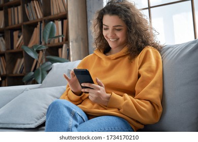 Счастливая латиноамериканская девочка-подросток проверяет социальные сети, держа смартфон дома. Улыбающаяся молодая латинская женщина с помощью мобильного телефона, играя в игру, делая покупки в Интернете, заказывая доставку, отдыхайте на диване