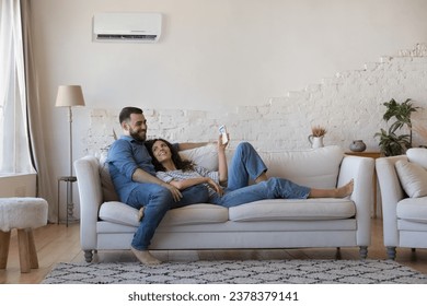 Happy millennial husband and wife resting on couch at home, relaxing under cooling air from conditioner, holding remote control for AC, using domestic appliance for comfort