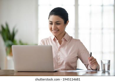 Happy Millennial Indian Businesswoman Sit At Desk In Office Work Online On Laptop Busy With Paperwork. Smiling Young Mixed Race Female Employee Worker Look At Computer Screen Consult Client Distant.