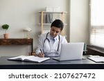 Happy millennial Indian female medical worker doctor in white uniform making notes in registration journal, managing appointments or prescribing illness treatment for patient, working on laptop.