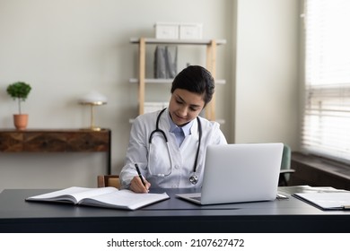 Happy millennial Indian female medical worker doctor in white uniform making notes in registration journal, managing appointments or prescribing illness treatment for patient, working on laptop.