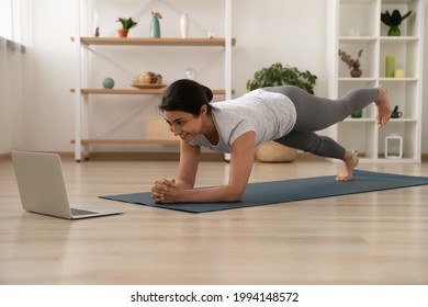 Happy millennial Indian female in sportswear on mat at home practice yoga stretching with online lesson or class on computer. Smiling ethnic woman train do workout watch video session on laptop. - Powered by Shutterstock