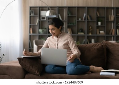 Happy Millennial Indian Female Student Sit On Sofa At Home Study Online On Laptop Take Notes In Notebook. Smiling Young Mixed Race Woman In Earphones Write Watch Webinar Work Distant On Computer.