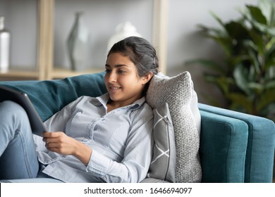 Happy millennial Indian girl lying on comfortable couch in living room watching movie on tablet, smiling young ethnic woman relax rest on cozy sofa furniture at home using modern pad gadget - Powered by Shutterstock