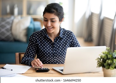 Happy millennial Indian girl sit at desk in living room manage household finances make payments on Internet on laptop, smiling young ethnic woman calculate expenditures pay bills on computer online