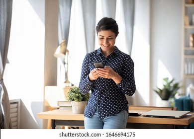 Happy millennial Indian girl stand in living room have fun texting messaging on modern smartphone gadget, smiling young ethnic woman using cellphone, shopping online or browsing Internet on device