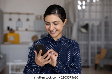 Happy millennial Indian girl, young woman reading text message on smartphone screen, smiling, feeling joy, chatting, typing, using online service, virtual app for payment on internet at home