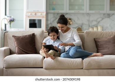 Happy Millennial Indian Mom Gen Z Son Boy Reading Book, Playing Videogame On Tablet Computer, Using Online App, Service, Chatting. Mother And Kid Shopping, Booking On Internet At Home