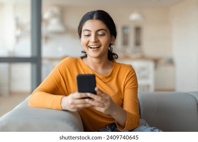 Happy millennial indian woman relaxing on comfortable couch, holding smartphone in hands, smiling young eastern lady chatting in social networks, watching videos, using mobile applications at home - Powered by Shutterstock